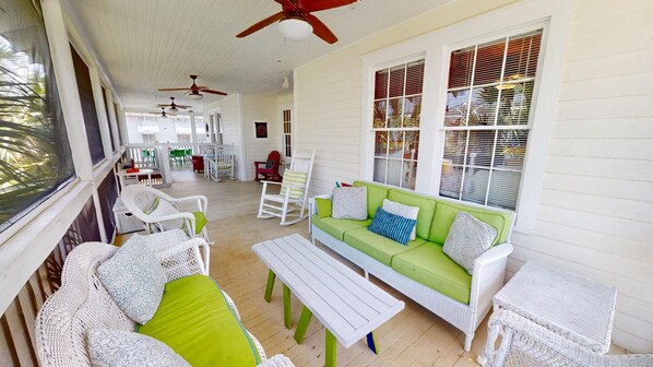 A covered porch with a white wicker sofa, green cushions, and a white table. There are ceiling fans, a rocking chair, and a dining area in the background.
