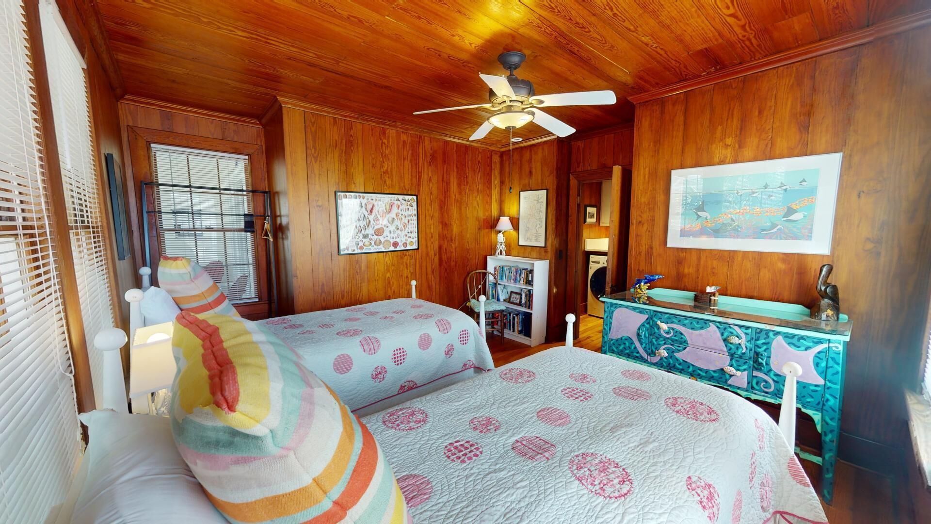 Un dormitorio acogedor con dos camas, paredes de madera, un ventilador de techo, una estantería y un mueble decorativo.
