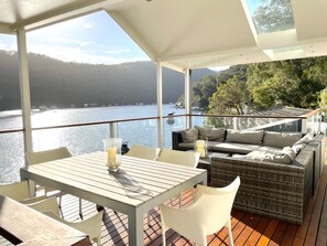 A deck with a table and chairs, a sofa, and a view of a lake and mountains. - Waterfront modern property w/ boat, outside hot tub, SUPs, kayaks, fishing equip (Berowra Creek)
