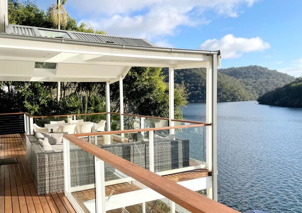 Una terraza moderna al aire libre con barandilla de vidrio, muebles blancos y vista a un lago rodeado de follaje.