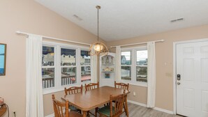 A dining room with a wooden table and chairs, large windows with white curtains, a chandelier, and a white door. - Fun-N-The Sun - 2 BR Home; Dog Friendly with an Ocean View! (Surf City)