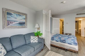 Um quarto com um sofá cinzento, uma cama com colcha estampada, um quadro na parede e um vaso de planta.