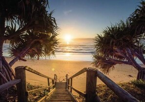 Eine Holztreppe führt zu einem Strand, an beiden Seiten Palmen.