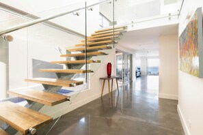 Un interior moderno con una escalera de cristal, un jarrón rojo sobre una mesita y vista al mar a través de un ventanal.