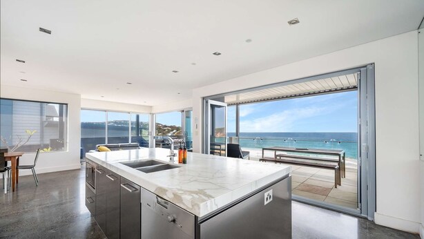 A modern kitchen with a large island, a dining area with a bench, and a view of the ocean through large sliding doors. - “Oceanview & Seabreeze” - with Pool (Copacabana)