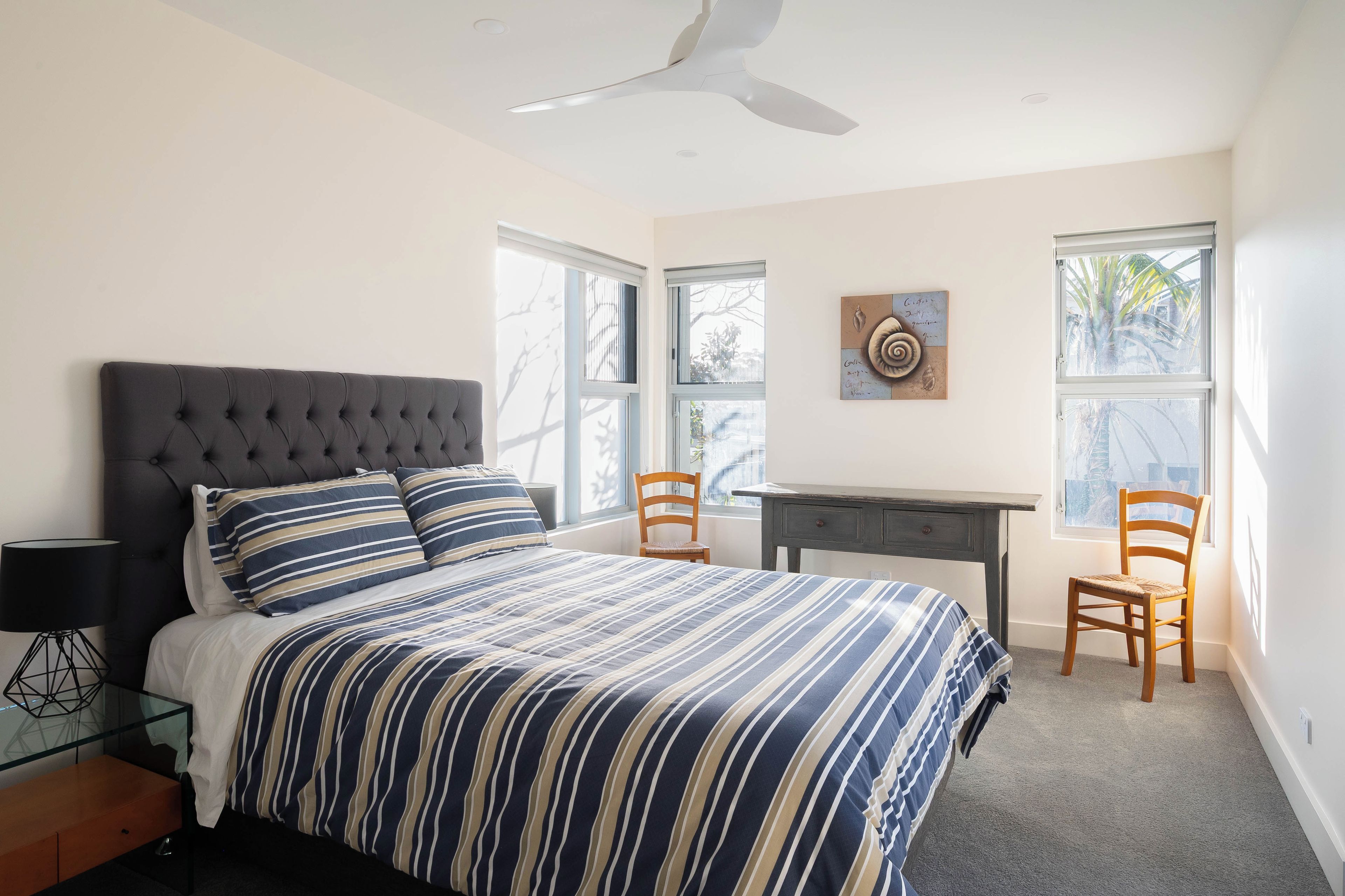 A bedroom with a tufted headboard bed, striped bedding, a desk, two chairs, and a wall clock.