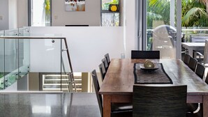 A modern kitchen with a glass staircase, a dining area with a wooden table and chairs, and a view of palm trees through large windows. - “Oceanview & Seabreeze” - with Pool (Copacabana)
