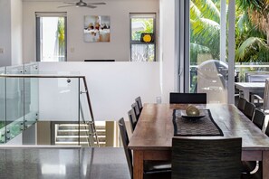 A modern kitchen with a glass staircase, a dining area with a wooden table and chairs, and a view of palm trees through large windows. - “Oceanview & Seabreeze” - with Pool (Copacabana)