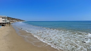 Une plage avec un ciel bleu dégagé, un rivage sablonneux et un bâtiment surmonté d’une tour de surveillance pour les sauveteurs.