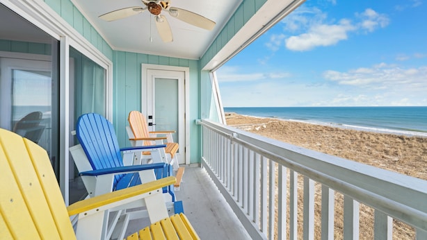 A balcony with a view of the beach, featuring a ceiling fan, a door, and several Adirondack chairs.
