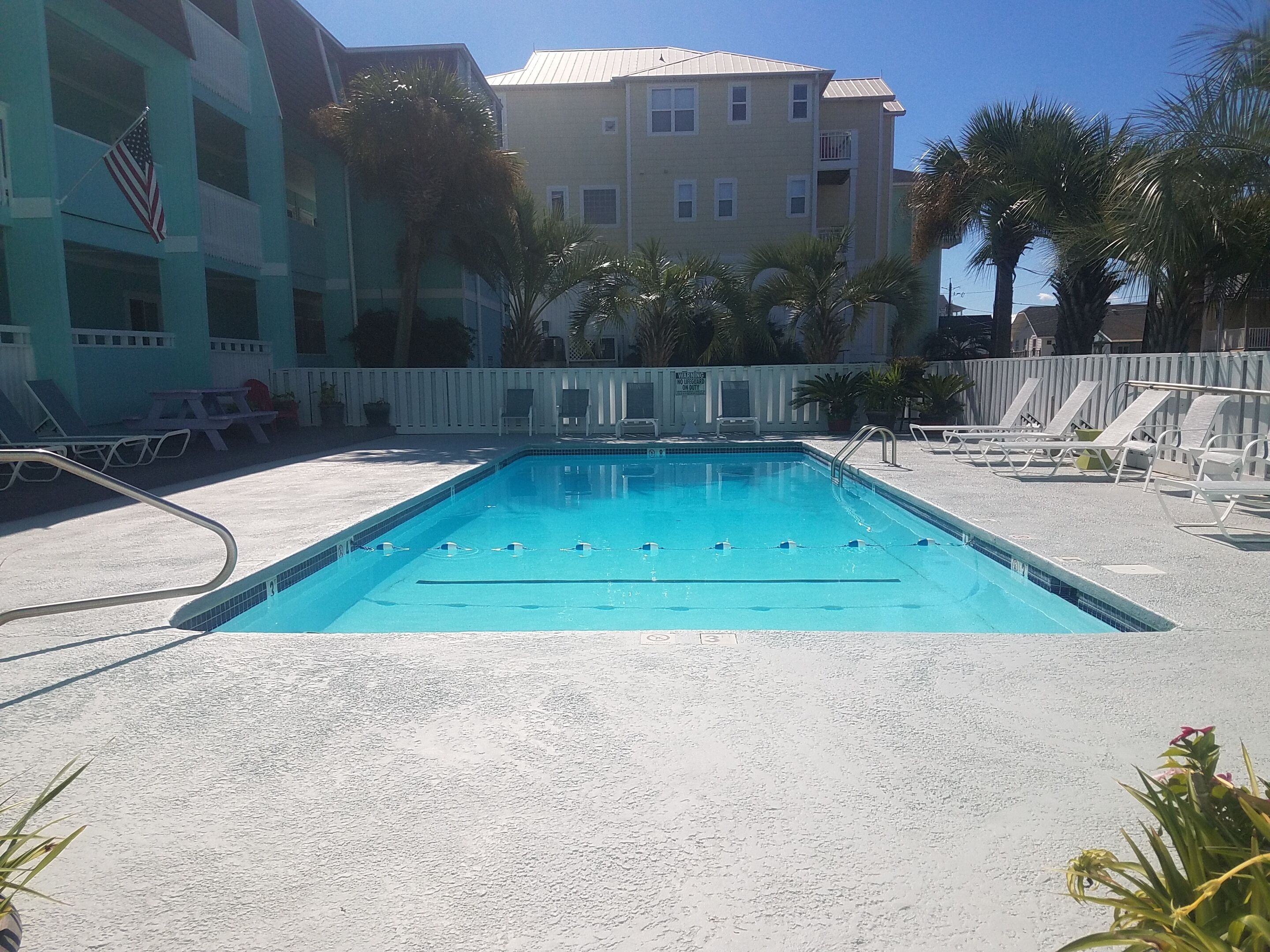 Ein Swimmingpool mit Liegestühlen und einer Flagge, umgeben von Palmen und einem Gebäude mit Balkonen.