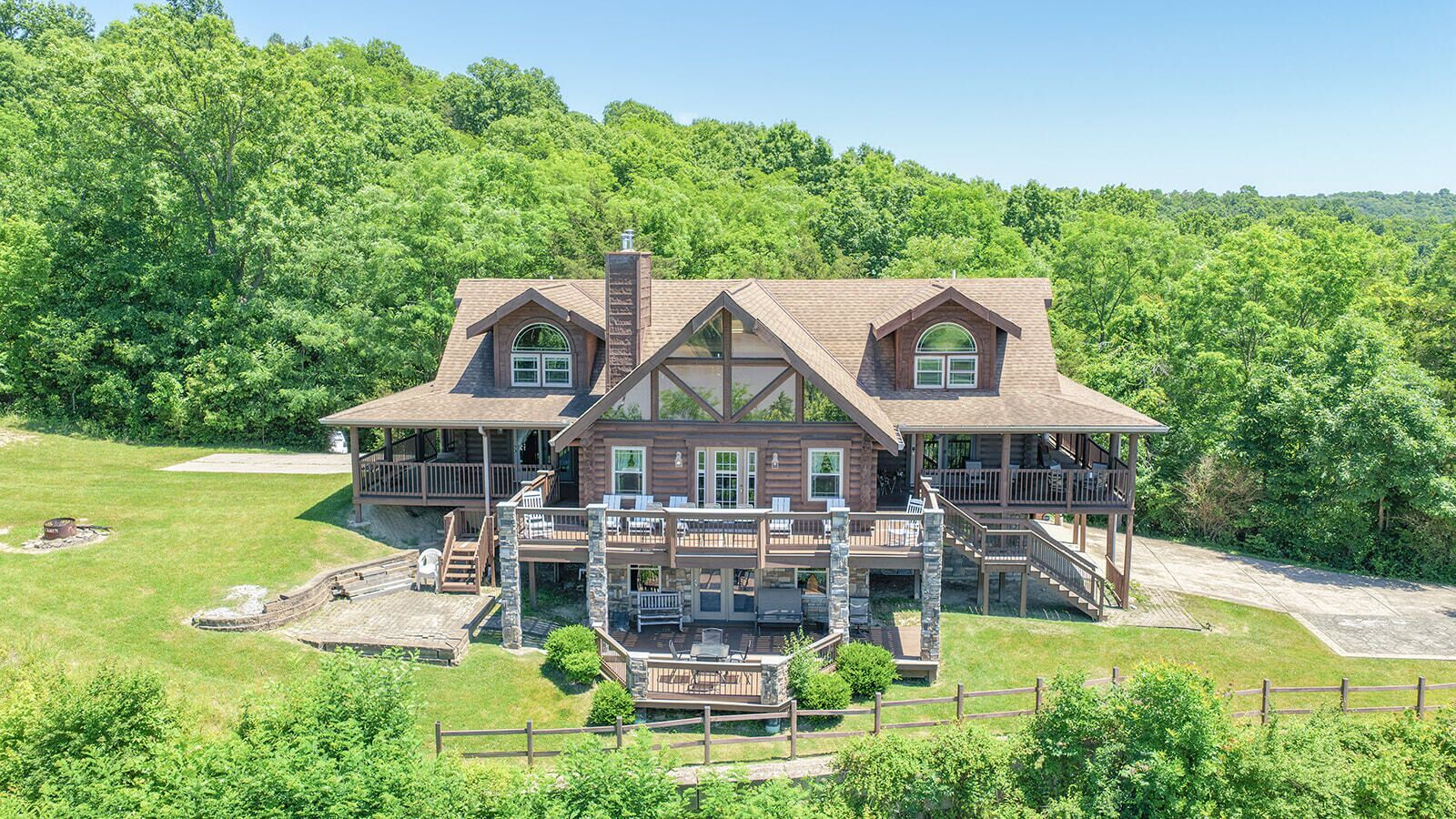 A large, two-story log cabin with multiple decks and a stone foundation, surrounded by lush greenery.