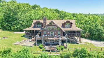 A large, two-story log cabin with multiple decks and a stone foundation, surrounded by lush greenery.
