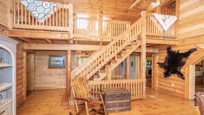A log cabin interior with a wooden staircase, a rocking chair, a chest of drawers, and a hanging black dog.