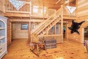 A log cabin interior with a wooden staircase, a rocking chair, a chest of drawers, and a hanging black dog.