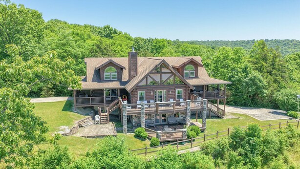 A large log cabin with a wrap-around porch, multiple decks, and a stone chimney.