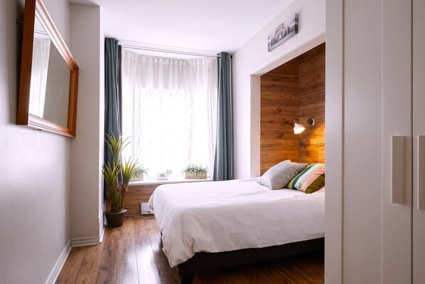A bedroom with a bed, a window with curtains, a potted plant, and a wooden accent wall. - MTLVR #11 | Central, Group Friendly, Patio (Montreal)