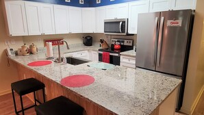 A kitchen with white cabinets, a stainless steel refrigerator, a microwave, and a red kettle. There are two black bar stools and a red placemat on the counter.