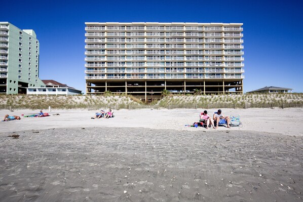 Un hotel frente a la playa con varios pisos, zonas de estar al aire libre y una playa de arena en primer plano.