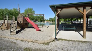 A playground with a red slide and climbing structures, a covered picnic area with benches, and a body of water in the background.