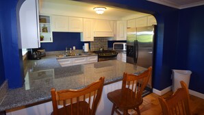 A kitchen with white cabinets, a granite countertop, a stainless steel refrigerator, and wooden chairs.