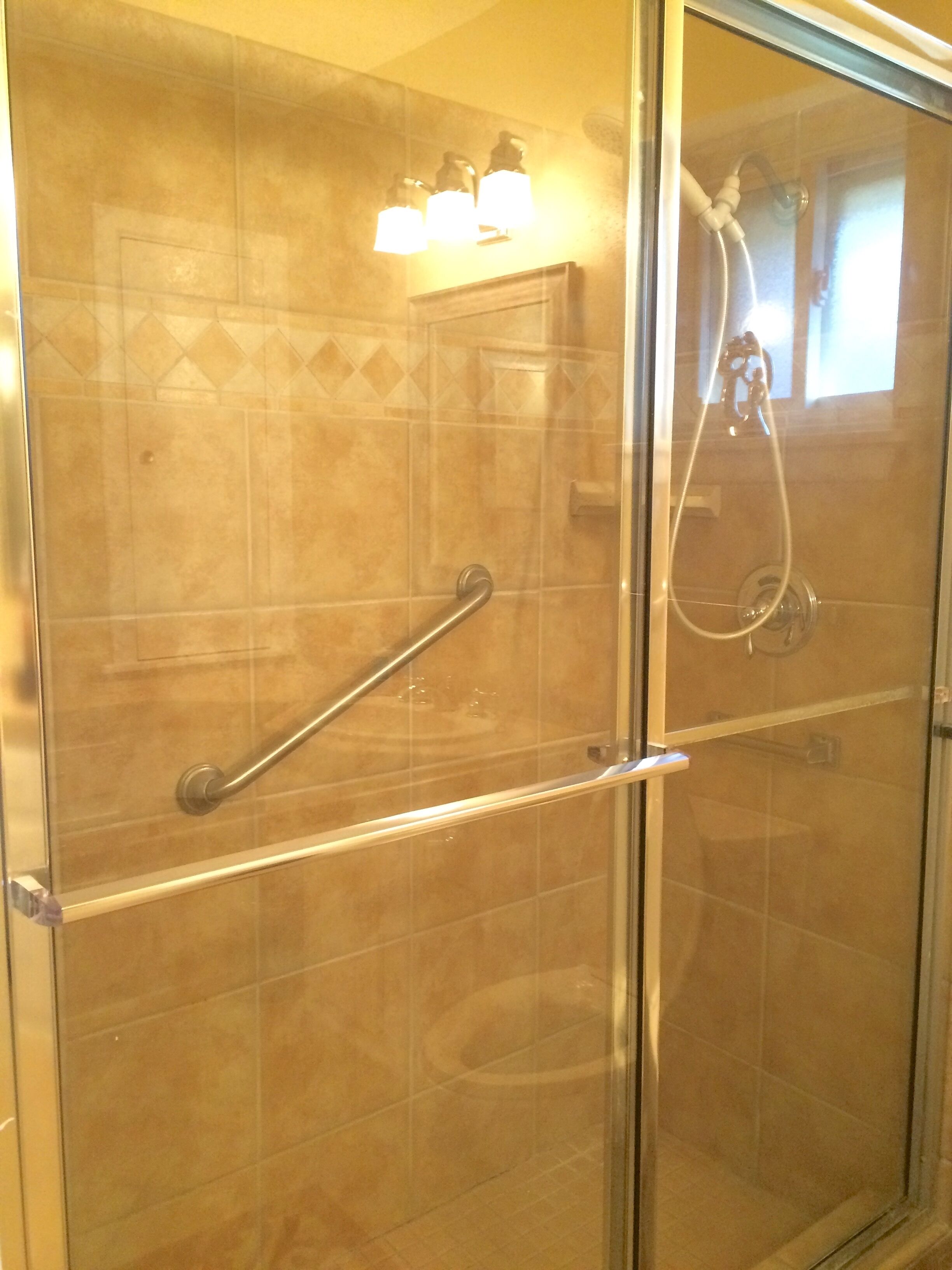 A glass-enclosed shower with a handrail, a handheld showerhead, and a wall-mounted light fixture.