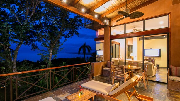 Un vaste balcon doté de mobilier en bois, d’un coin repas et offrant une vue sur l’océan au crépuscule.