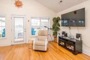 A living room with a beige armchair, a television mounted on the wall, a wooden floor, and a large window with blinds. - Gone Coastal - 2nd Row home; 4BR; Ocean Views from Sun Deck (Surf City)