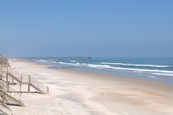 Een strand met een houten trap die naar het zand leidt, een pier die in het water steekt, en een heldere lucht.