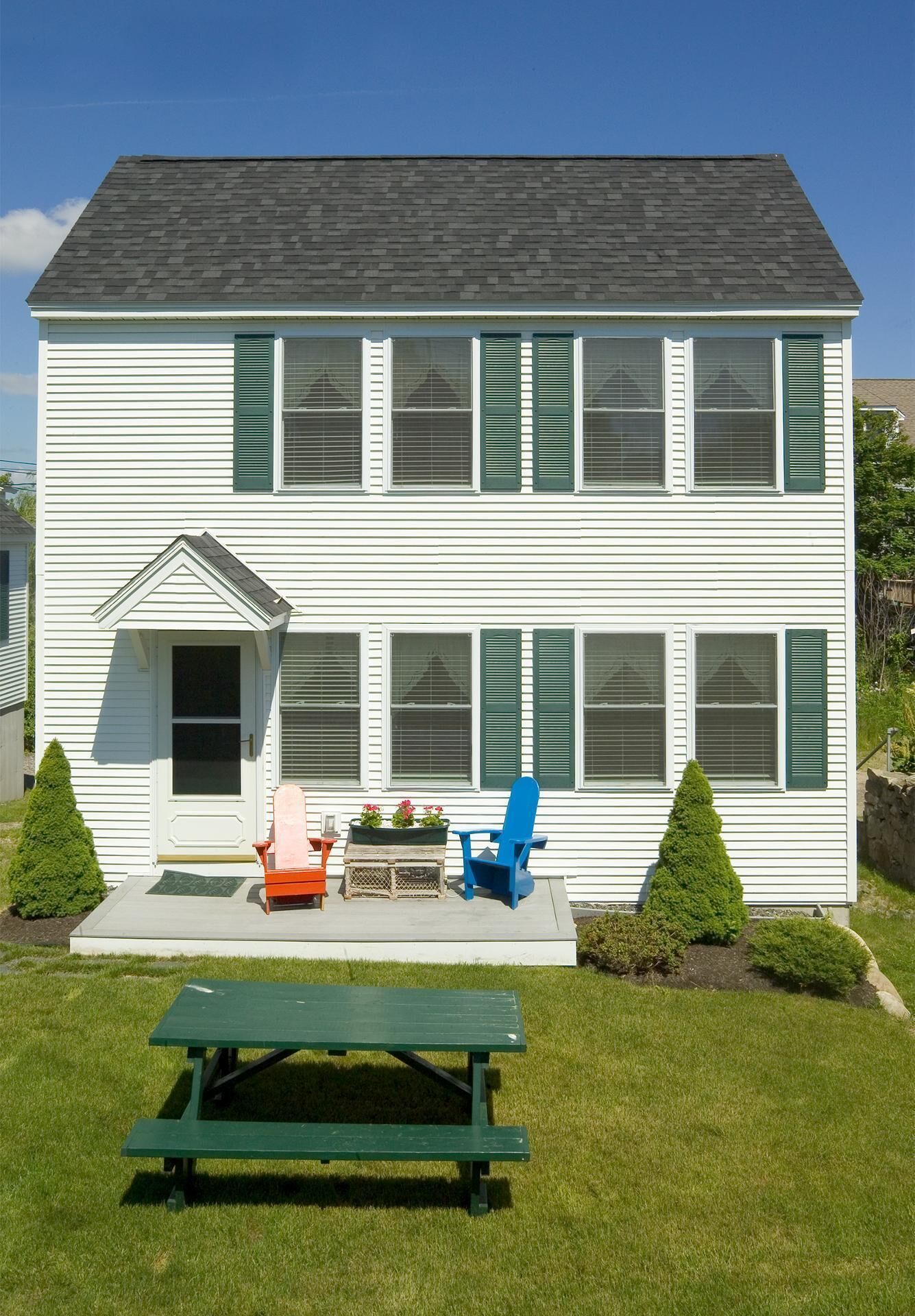 A two-story white house with green shutters, a front porch with a bench, and a picnic table in the front yard.