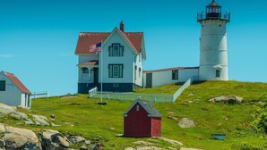 A lighthouse with a white exterior and a red roof, a white house with a red roof, and a small red shed. - Goose Rocks Cottage at One Long Beach (York Beach)