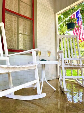 A porch with white rocking chairs, a small table with two glasses, and an American flag.