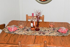 A wooden dining table with a placemat, two wine glasses, a bottle of wine, and a decorative vase. - Old Country Charm 2 1/2 blocks from Main St Shopping/Private Get-a-Way w/Hot Tub (Fredericksburg)