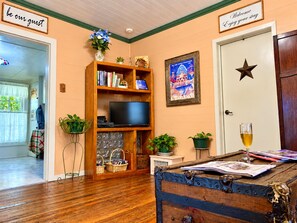 A cozy room with a wooden bookshelf, a flat-screen TV, a vintage suitcase, and a glass of wine on a wooden table.