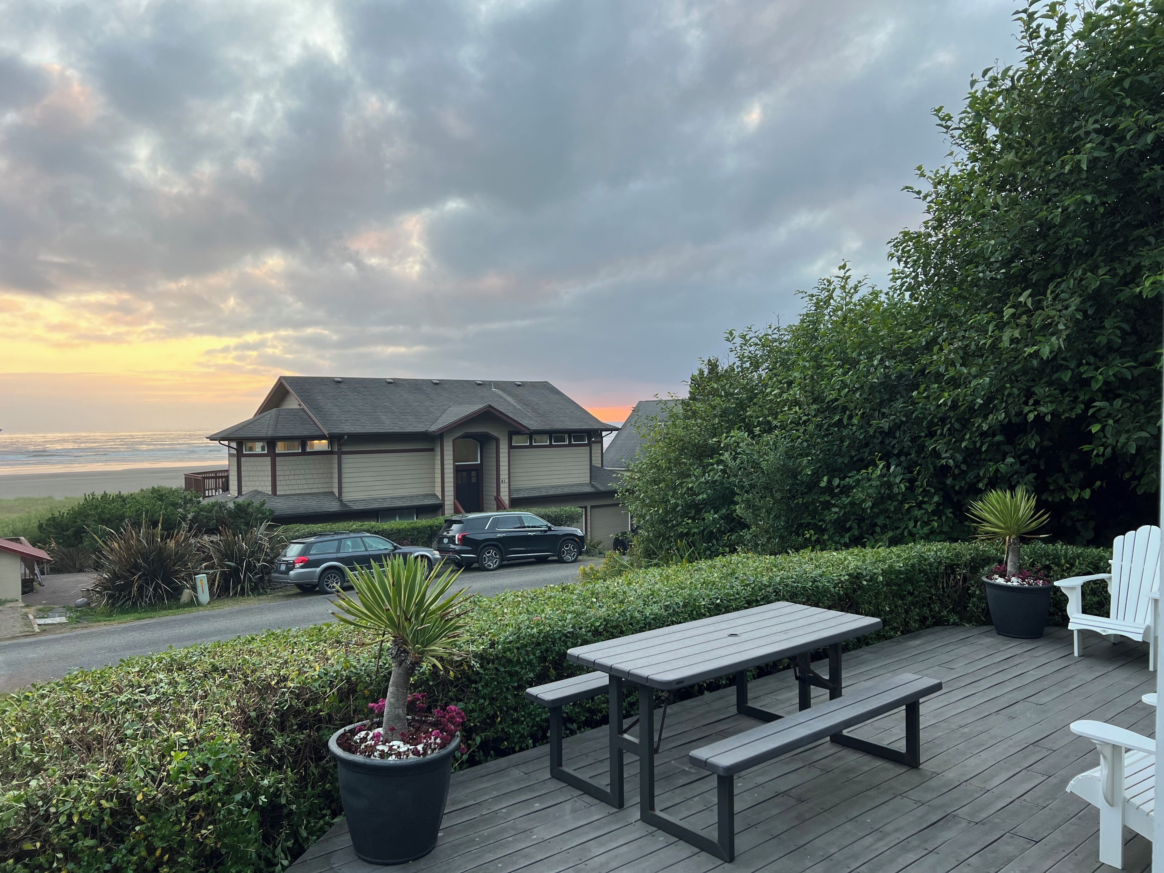A deck with picnic tables overlooking a beach and a house with parked cars.