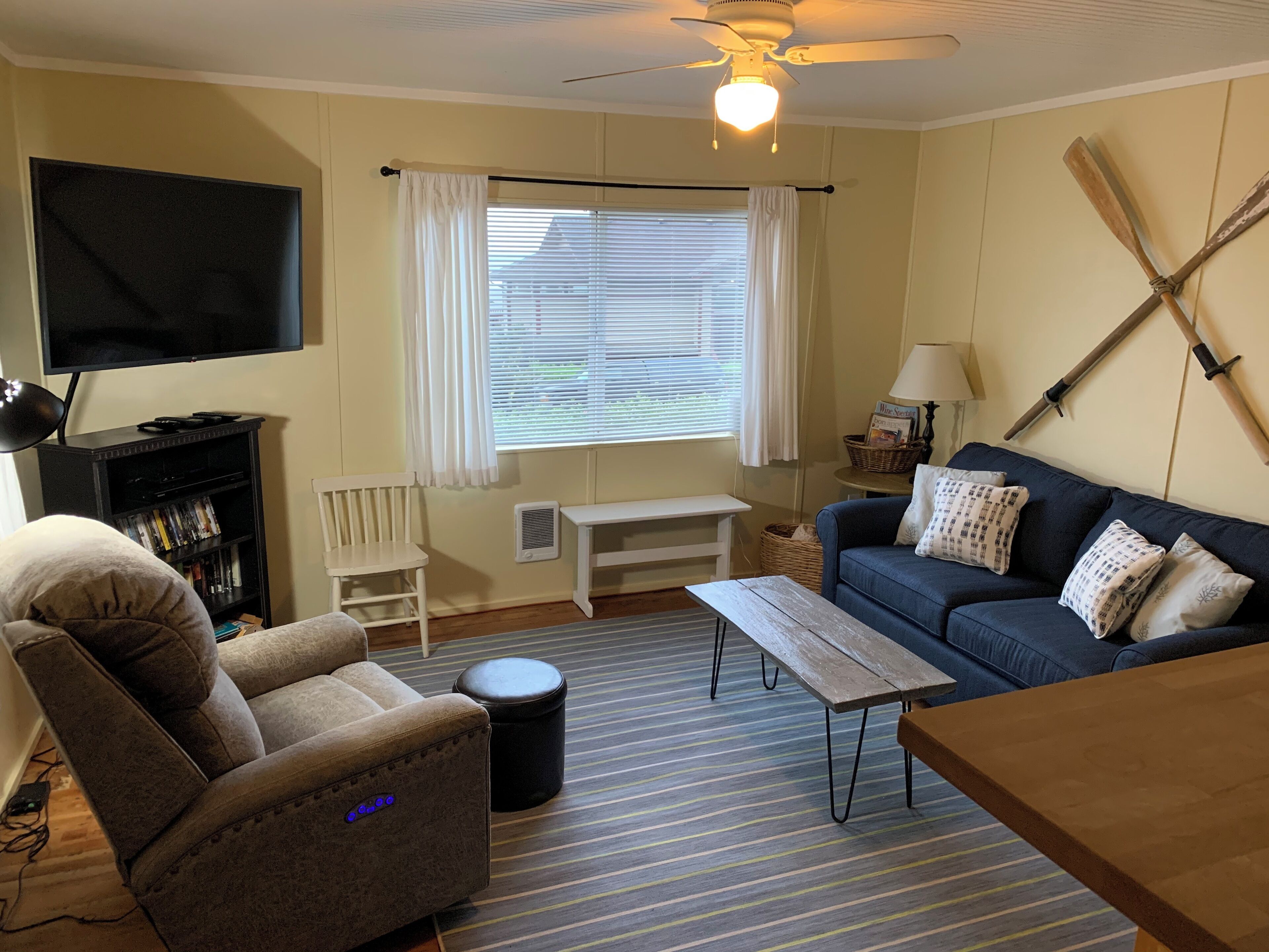 Ein Wohnzimmer mit einer Couch, einem Sessel, einem Fernseher, einem Deckenventilator und einem Fenster mit Vorhängen.