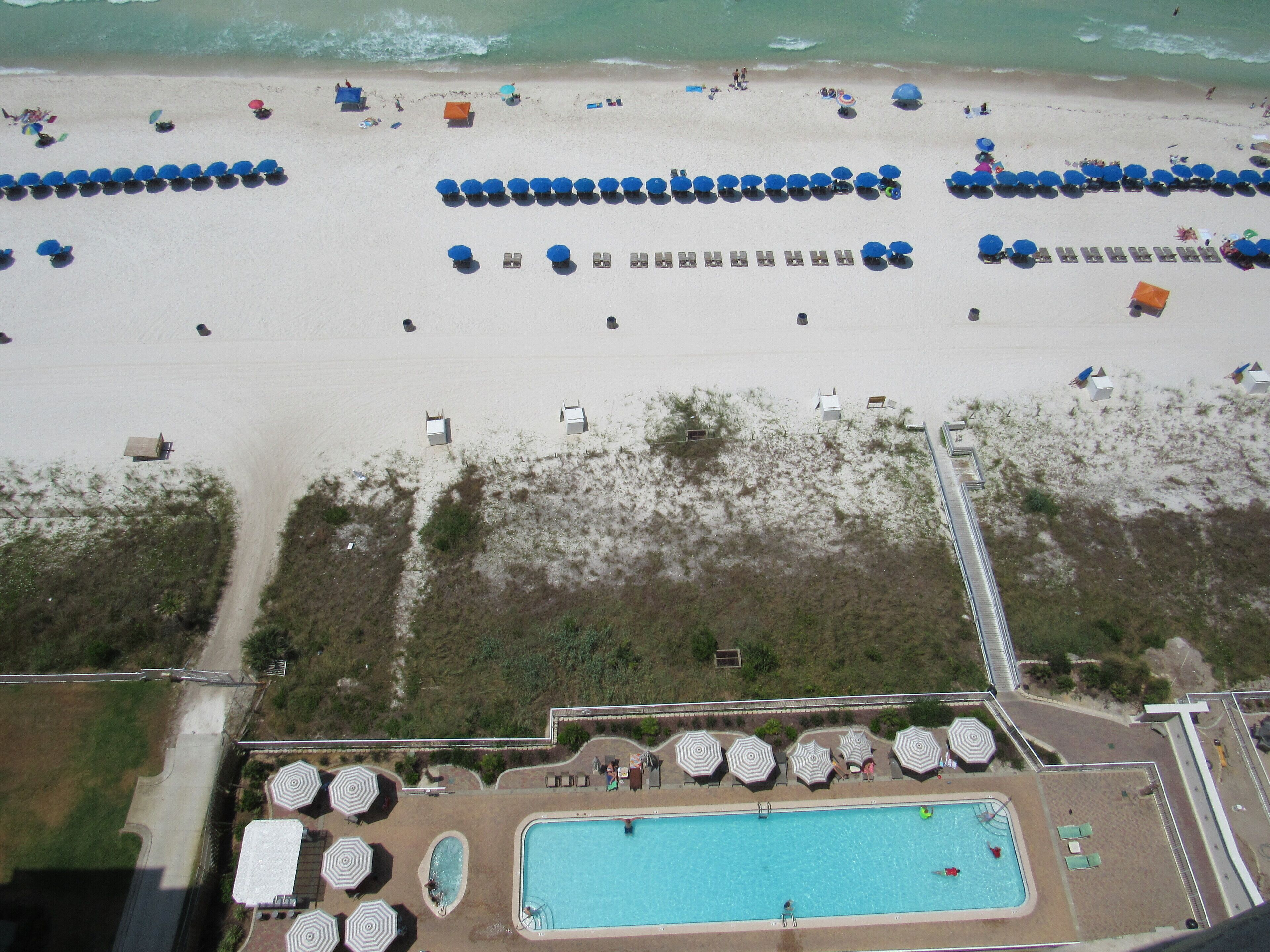An aerial view of a beach with rows of umbrellas and lounge chairs, a swimming pool, and a road leading to the beach.