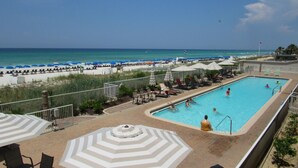 Une piscine avec des chaises longues et des parasols, une plage en arrière-plan et un ciel dégagé.