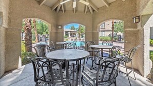 Un patio al aire libre con mesas y sillas redondas, un ventilador de techo y vista a una piscina y edificios al fondo.