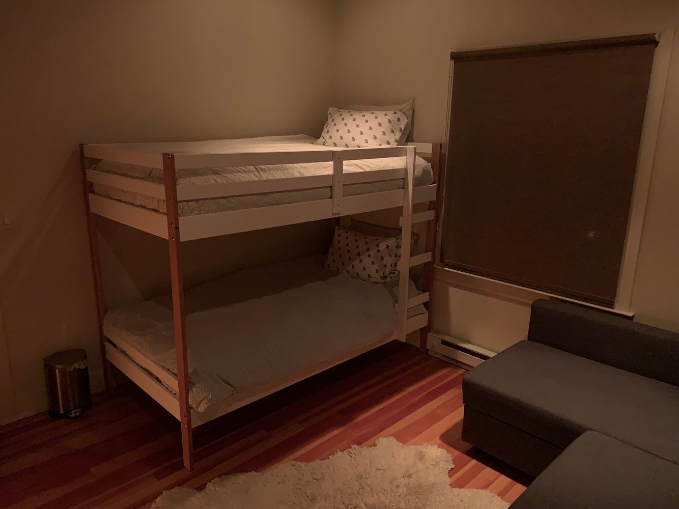 Een stapelbed met matras en kussens, een kleine prullenbak, een grijze bank en een raam met verduistering.