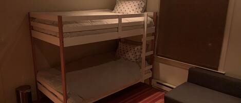 Een stapelbed met matras en kussens, een kleine prullenbak, een grijze bank en een raam met verduistering.