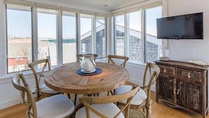 Una mesa de madera redonda con seis sillas, un televisor montado en la pared y ventanas grandes que dan vista a edificios y una masa de agua.