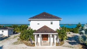 A two-story house with a central entrance and a small porch, surrounded by palm trees and a view of the ocean. - Escape to Exhale at a modern style villa with breathtaking views of chalk sound (Providenciales)