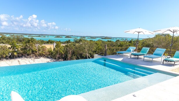 A clear blue pool with lounge chairs and umbrellas, surrounded by tropical vegetation and a view of the ocean. - Escape to Exhale at a modern style villa with breathtaking views of chalk sound (Providenciales)