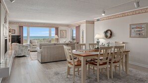 A spacious living room with a large window overlooking the ocean, a dining area with wooden chairs, and a cozy seating area with a fireplace. - DS203 Ocean-Front Condo with Pool and Ocean Views! (Nags Head)