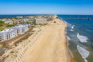 Una zona costera con una playa de arena, un muelle que se adentra en el agua y edificios residenciales a lo largo de la costa.
