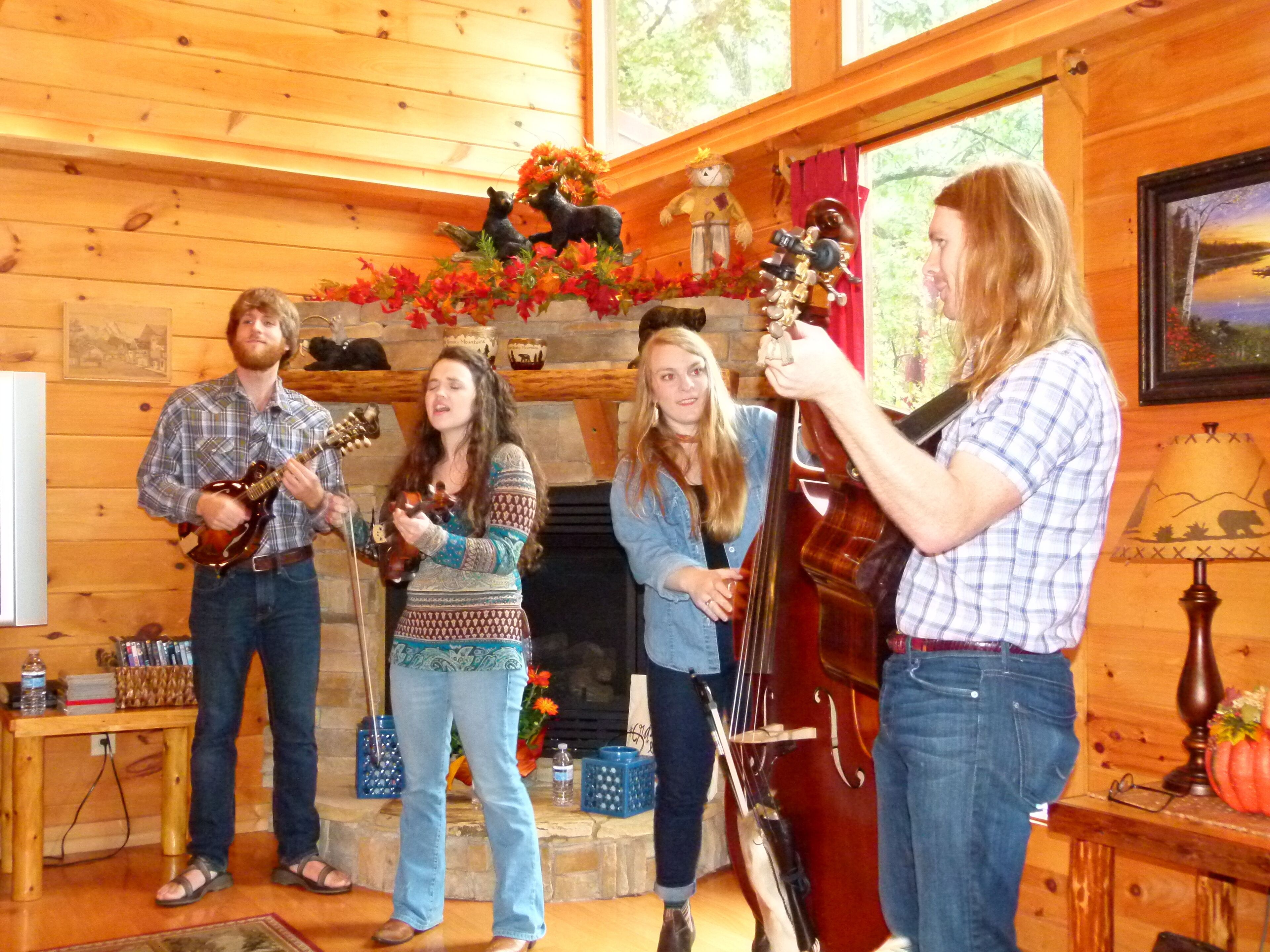 A group of people playing string instruments in a wooden room with a fireplace, a table with items on it, and a lamp.