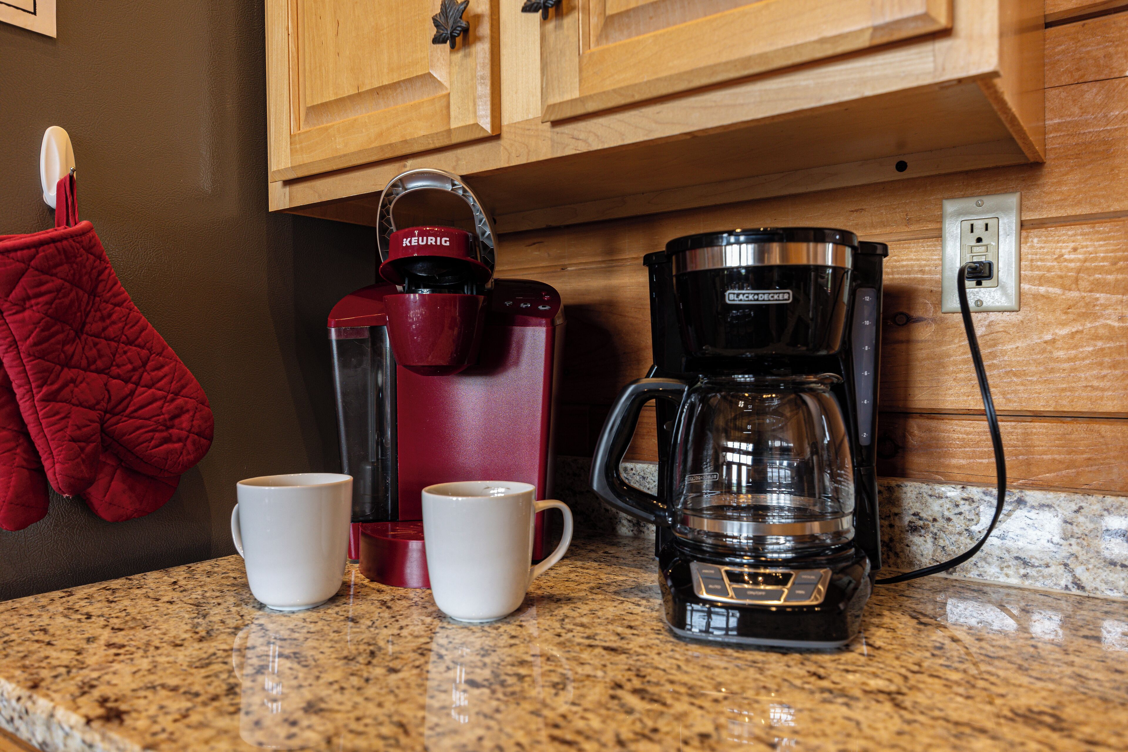 Una encimera de cocina con una cafetera, dos tazas y un hervidor rojo.