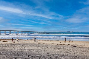 Una playa con un muelle que se adentra en el océano, arena y gente caminando.
