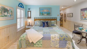 A bedroom with a large bed, a nightstand, a window with blinds, a wall-mounted shelf, and a painting.
